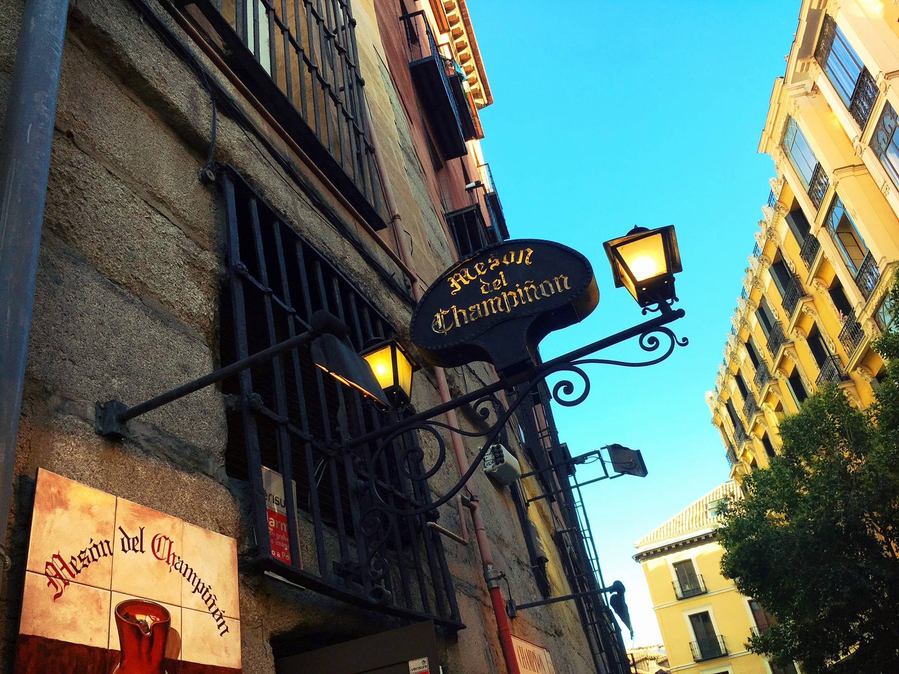 Letrero de forja del Mesón del Champiñón con un farol encendido en la fachada de la Cava de San Miguel